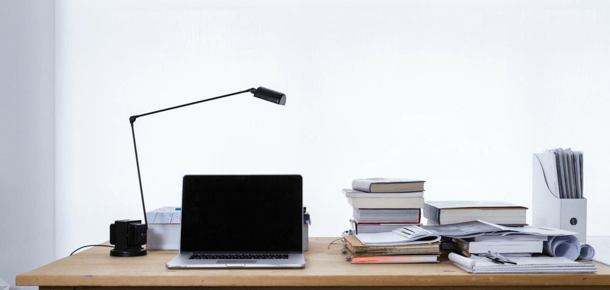 Laptop on table with lamp to left and stacks of books to right.